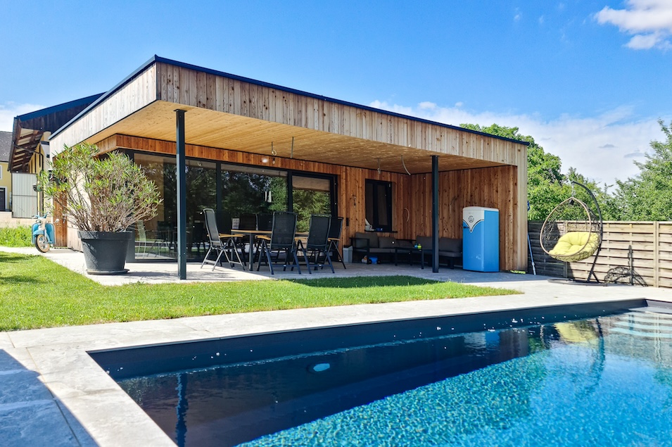 Pool mit frischem klaren Wasser und dahinter ein Poolhaus aus Holz mit Sitzgarnitur darunter.