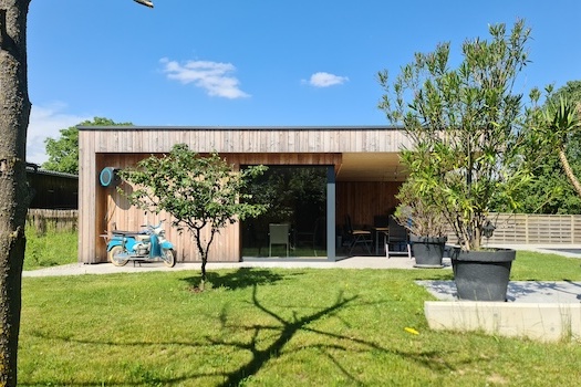 Seitlicher Blick auf das Poolhaus mit Holzfassade, davor im Garten stehen Bäume und Grünplfanzen. Vor dem Poolhaus steht ein blaues Moped.